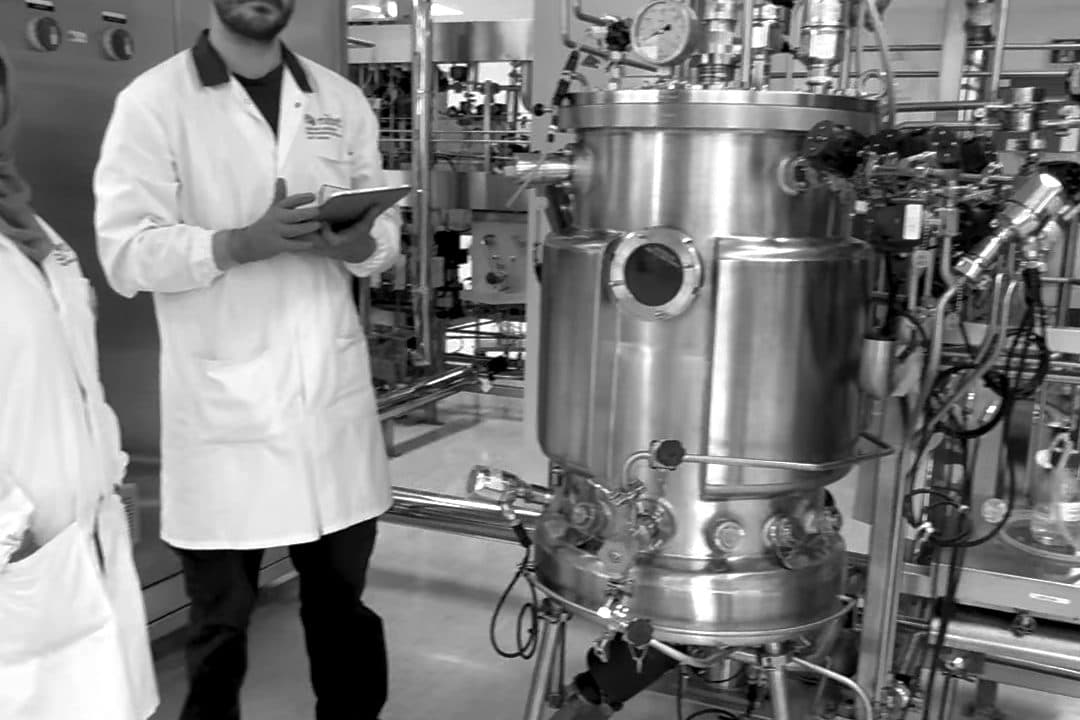 Two learners in lab coats, one holding an iPad, beside the bioreactor in NIBRT's upstream suite